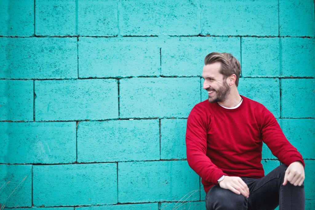 Mann mit rotem Pullover, lachend vor blauer Mauer

